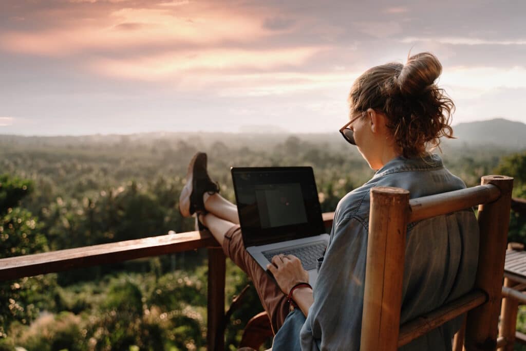 Junge Frau mit Laptop sitzt auf einer Holzveranda auf einer Anhöhe mit Blick auf ein großes Waldgebiet.