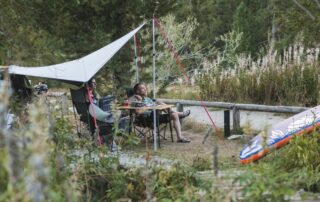 3 Freunde sitzen um einen Campingtisch unter einem Dachzelt vor dem Auto im Sommer in der natur