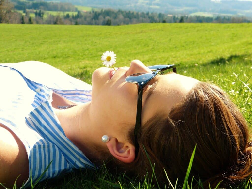 Frau liegt im Gras bei Sonnenschein mit Sonnenbrille und Gänseblümchen im Mund. Ausschnitt Kopf