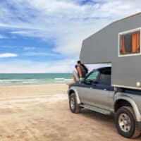 Pickup mit Campingaufsatz am Sandstrand vor türkisblauem Meer. Eine Frau sitzt auf der Motorhaube