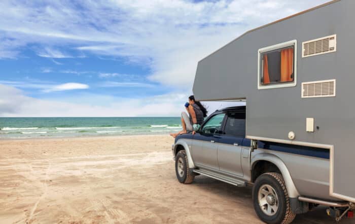 Pickup mit Campingaufsatz am Sandstrand vor türkisblauem Meer. Eine Frau sitzt auf der Motorhaube