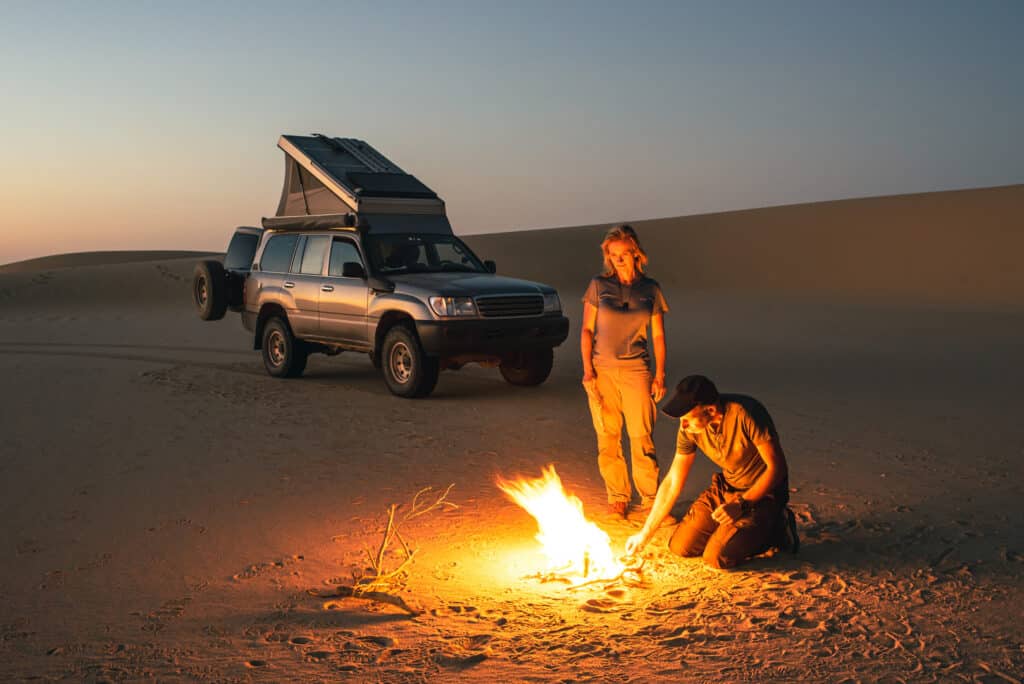 Pärchen in der Wüste am Abend am Lagerfeuer im Hintergrund ein Jeep mit Campingaufsatz