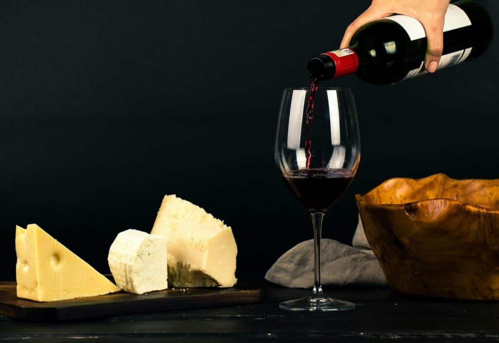 Glas, das gerade mit Rotwein aus einer Flasche befüllt wird steht auf einem dunklen Tisch daneben Käsestücke