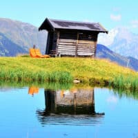 Heustadl aus Holz auf einer Wiese in den Bergen mit 2 Holzliegen vor einem See