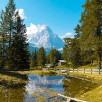 Blick über ein klares Gewässer das von Nadelbäumen gesäumt ist auf eine Bergspitze im Hintergrund bei blauem Himmel