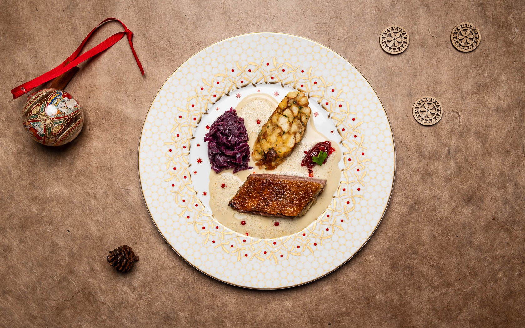 Teller mit einem knusprigen Stück Gänsebrust, Serviettenknödel, Blaukraut und Preiselbeeren auf schön dekorierte, weißem Teller auf goldener Tischdecke mit Weihnachtskugel, zwei Holzsternen und Tannenzapfen