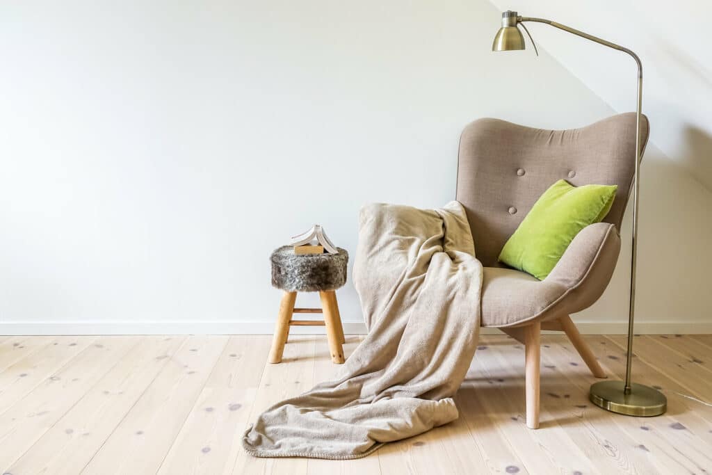 heller Lesesessel im skandinavischen Stil mit heller Wolldecke und hellgrü´nem Kissen daneben Stehlampe und Fellhocker als Tischchen mit Buch auf hellem Holzboden vor weißer Wand