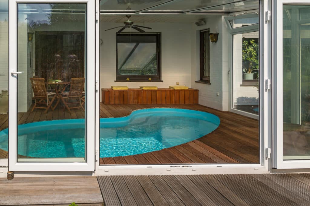 Wintergarten mit Holzboden und kleinem in den Boden eingelassenen Swimming-Pool. Im Hintergrund Tisch mit Stühlen und Holzbank mit gelben Kissen.