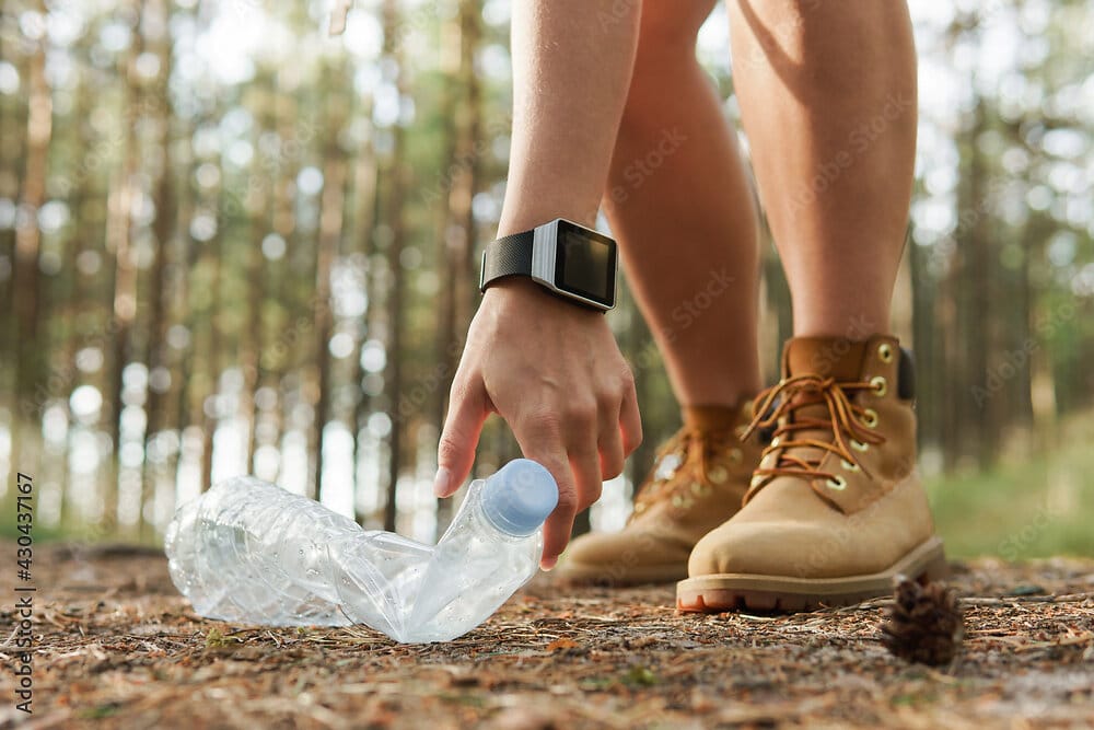 Ausschnitt Beine und Wanderschuhe einer jungen Frau in Shorts, die im Wald eine Plastikflasche aufliest.