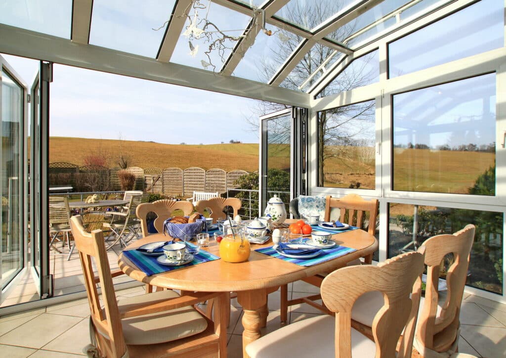 Ausblick aus einem Wintergarten am Feldrand mit geöffneten Türen und gedecktem Frühstückstisch.