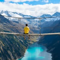 Junger Mann im gelben T-Shirt sitzt auf einer Hängebrücke über dem Schlegeisspeichersee vor Bergpanorama