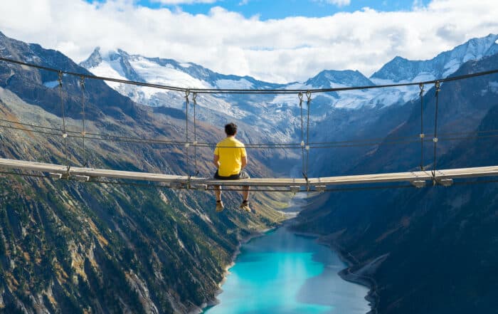 Junger Mann im gelben T-Shirt sitzt auf einer Hängebrücke über dem Schlegeisspeichersee vor Bergpanorama
