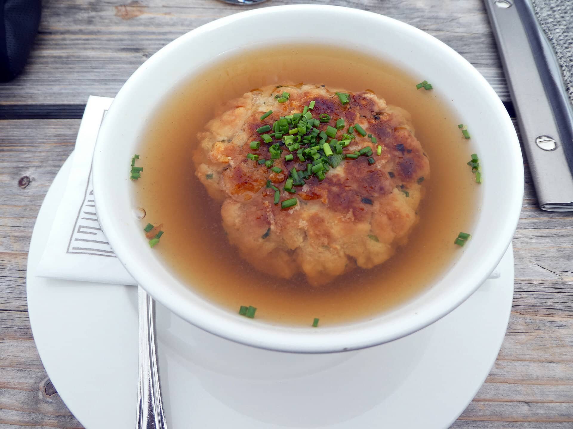 Tiroler Kaspressknödelsuppe in weißer Porzellanschüssel auuf Holztisch