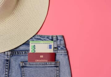 Auschnitt von Jeans und Strohhut auf rosafarbenem Hintergrund in der hinteren Tasche steckt ein Reisepass und ein 100 Euroschein