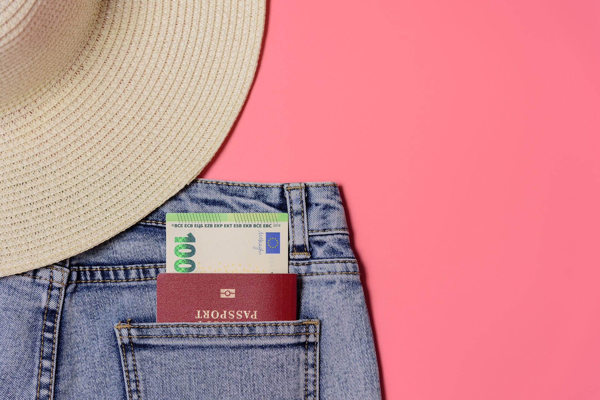 Auschnitt von Jeans und Strohhut auf rosafarbenem Hintergrund in der hinteren Tasche steckt ein Reisepass und ein 100 Euroschein
