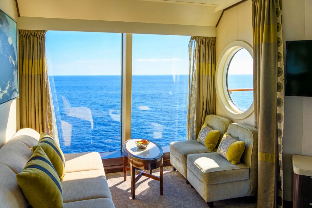 Luxuriöse Kabine auf Kreuzfahrtschiff mit gemütlicher Sofasitzgarnitur vor bodentiefen Fenstern mit Blick auf das tiefblaue Mittelmeer