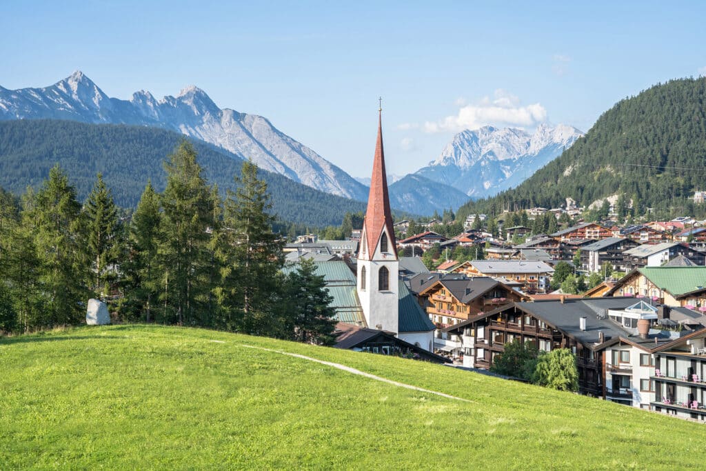 Blick auf Seefeld und mit Pfarrkirche. Im Vordergrund eine grüne Wiese im Hintergrund Berglandschaft im Sommer.