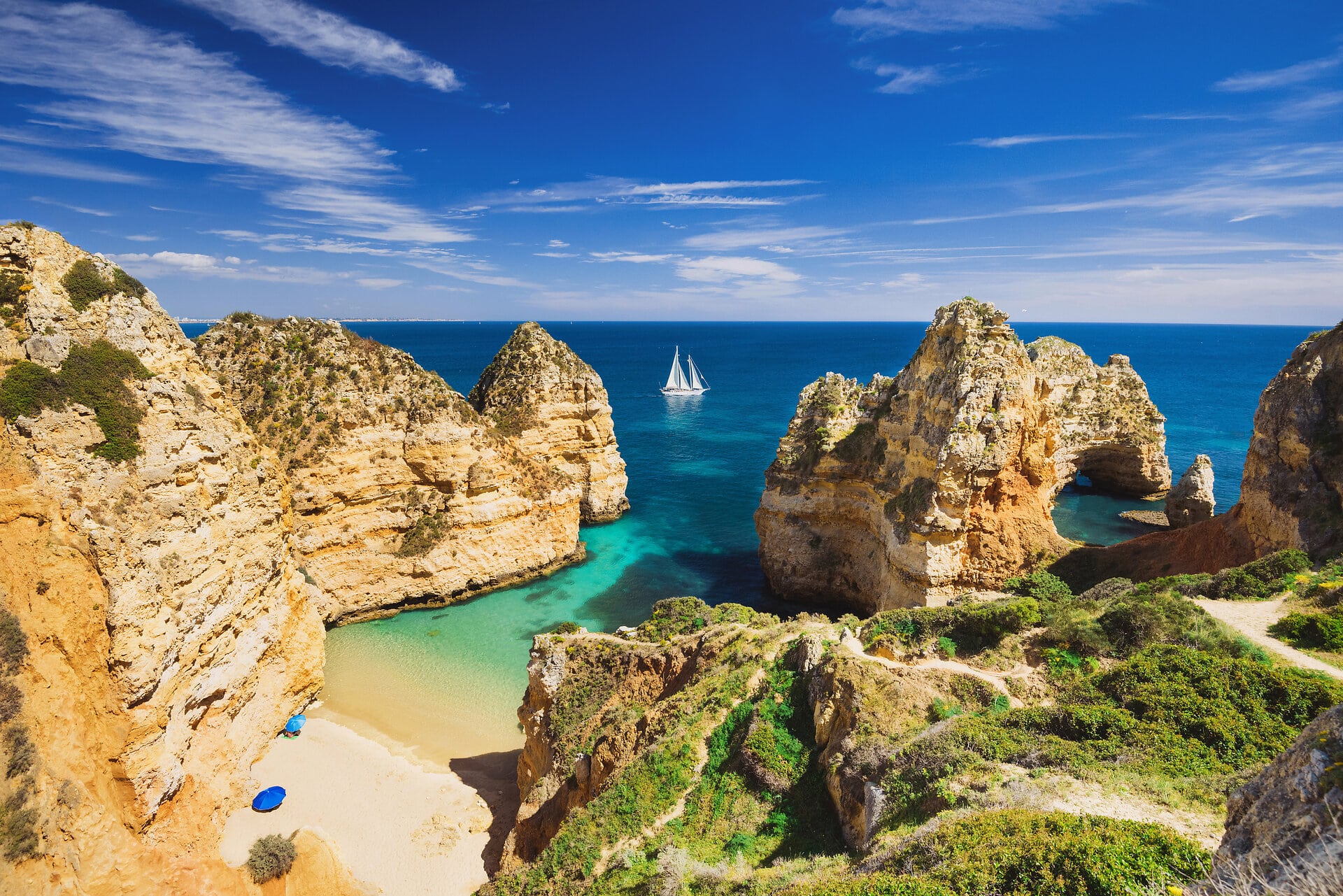 Blick über eine kleine von hohen Felsen gesäumte Bucht mit Sandstrand auf das türkisblau bis dunkelblau gefärbte Meer auf dem ein Segelboot fährt bei Sonnenschein