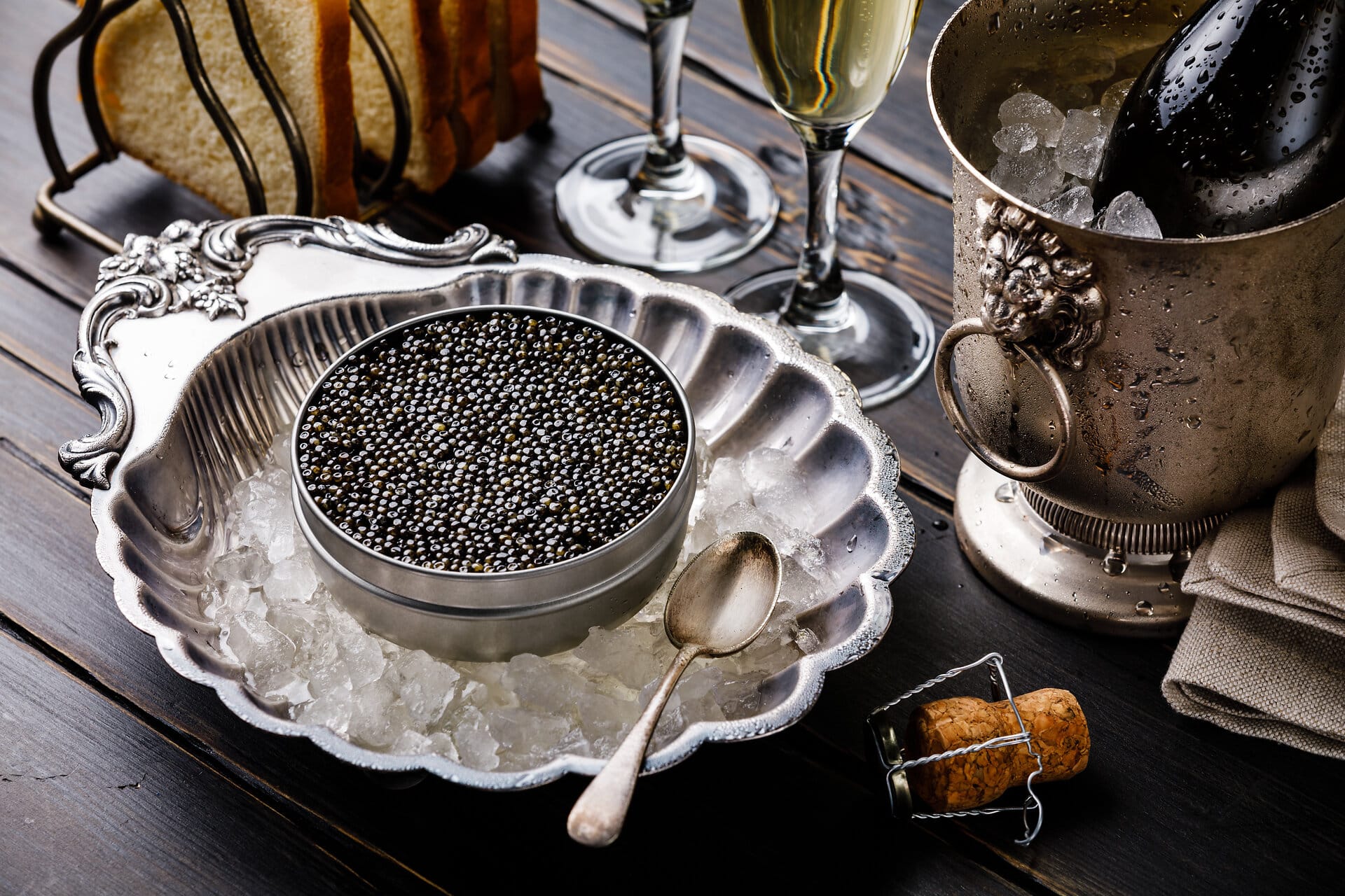 Black caviar in can on ice in silver bowl, bread and champagne in ice bucket on black wooden background