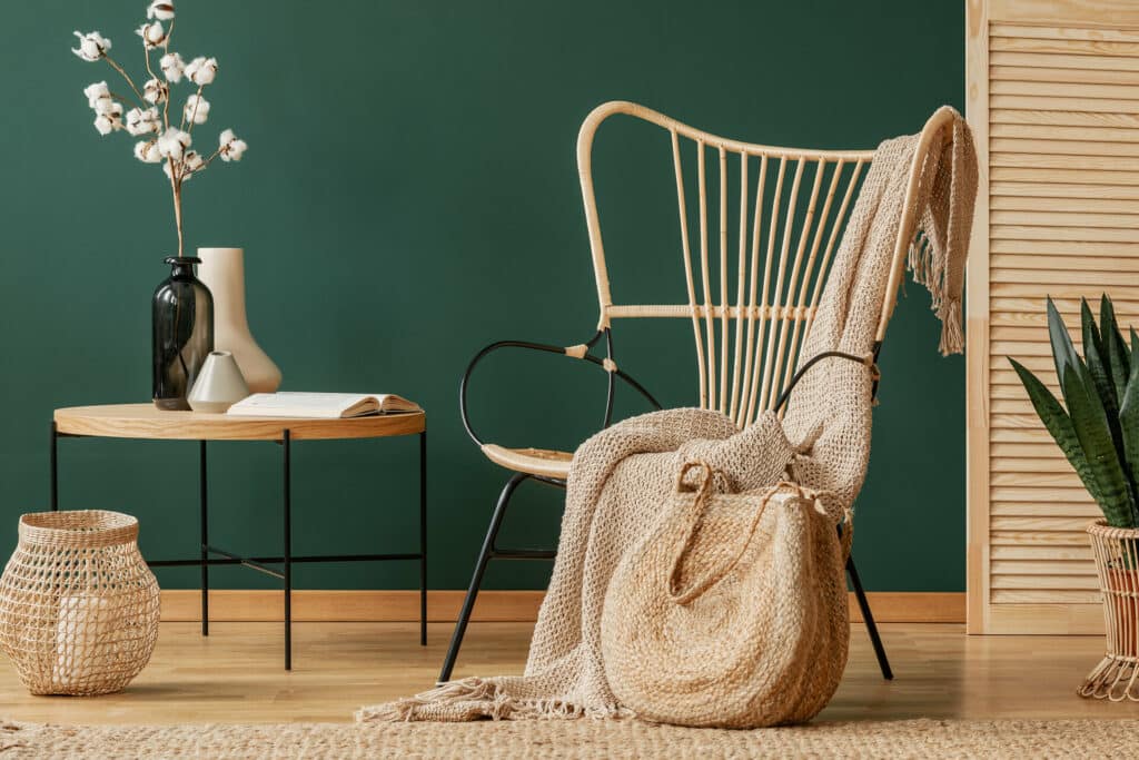 eleganter Rattanstuhl vor dunkelgrüner Wand mit Tisch und Vase mit weißen Orchideen und einer Decke und Tasche aus hellbeigen Naturmaterialien