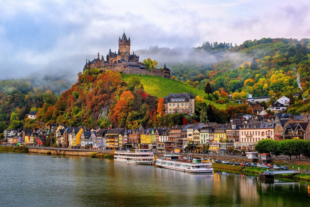 Blick vom Rhein aus auf Cochem und das Rheinufer mit 2 Ausflugsschiffen und auf Burg Cochem auf bunten herbstlichen Hügeln.