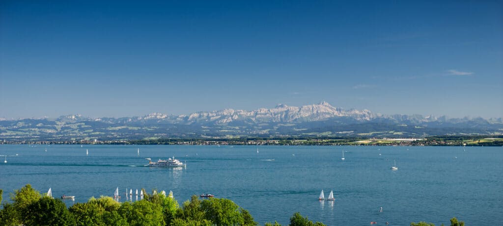 Blick über den sommerlichen Bodensee auf das Säntisgebirge in der Schweiz mit Segelbooten und Ausflugsschiff.