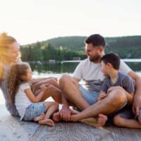 Glückliche Familie mit Junge und Mädchen sitzen in Shorts und T-Shirts barfuß auf einem Holzsteg an einem Seeufer im Sommer