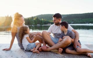 Glückliche Familie mit Junge und Mädchen sitzen in Shorts und T-Shirts barfuß auf einem Holzsteg an einem Seeufer im Sommer