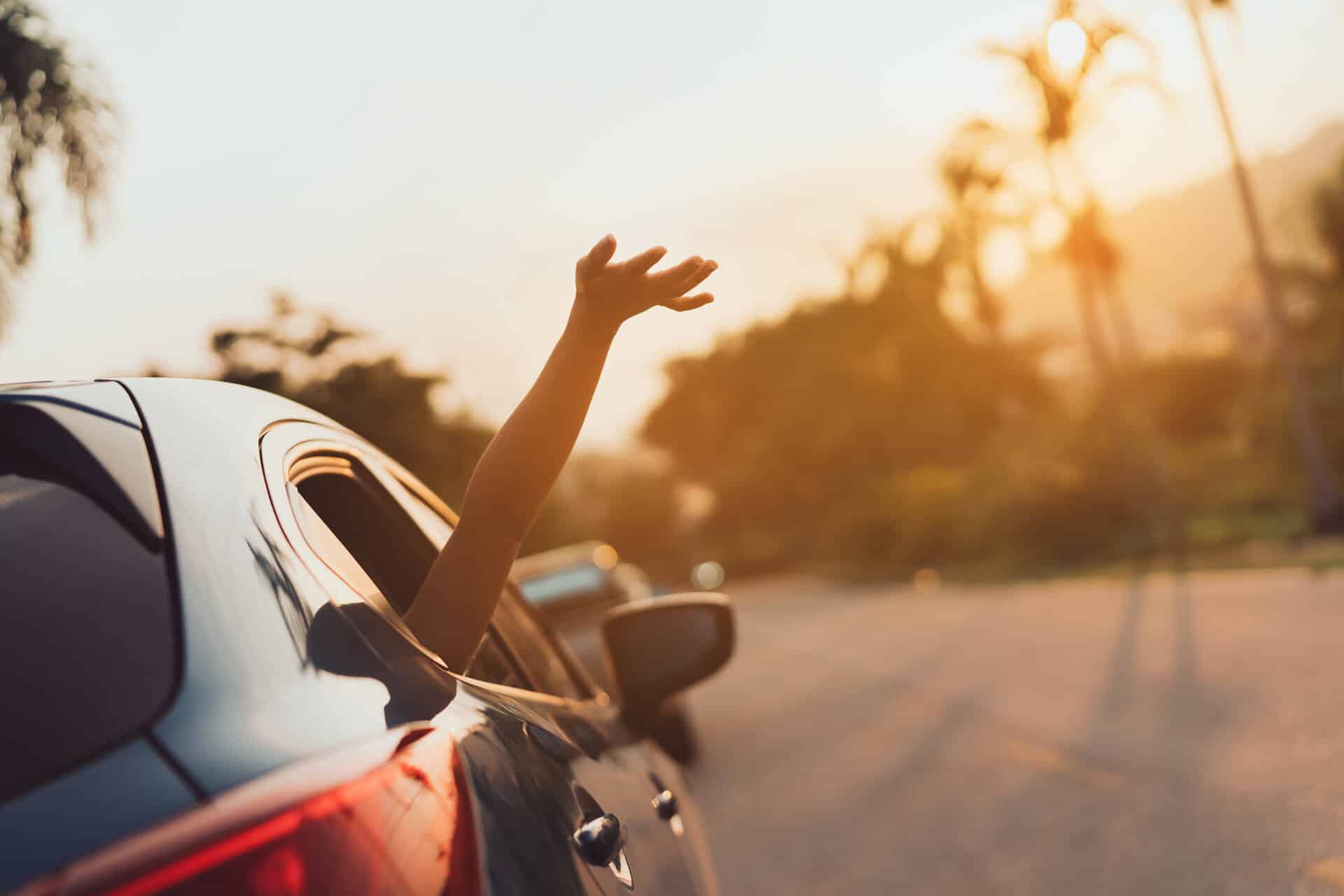 Auto im warmen Sonnenuntergang. Mädchenhand winkt aus dem hinteren Fenster. Urlaubsstimmung.