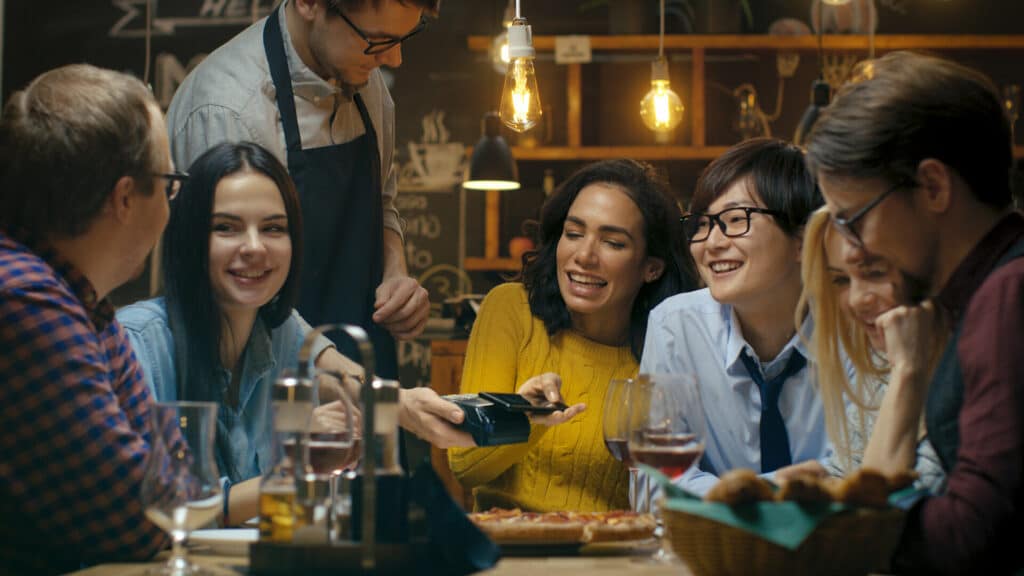 In the Bar Waiter Holds Credit Card Payment Machine and Beautiful Woman Pays for Her Order with Contactless Mobile Phone Payments System. She Has Good Time with Her Friends.