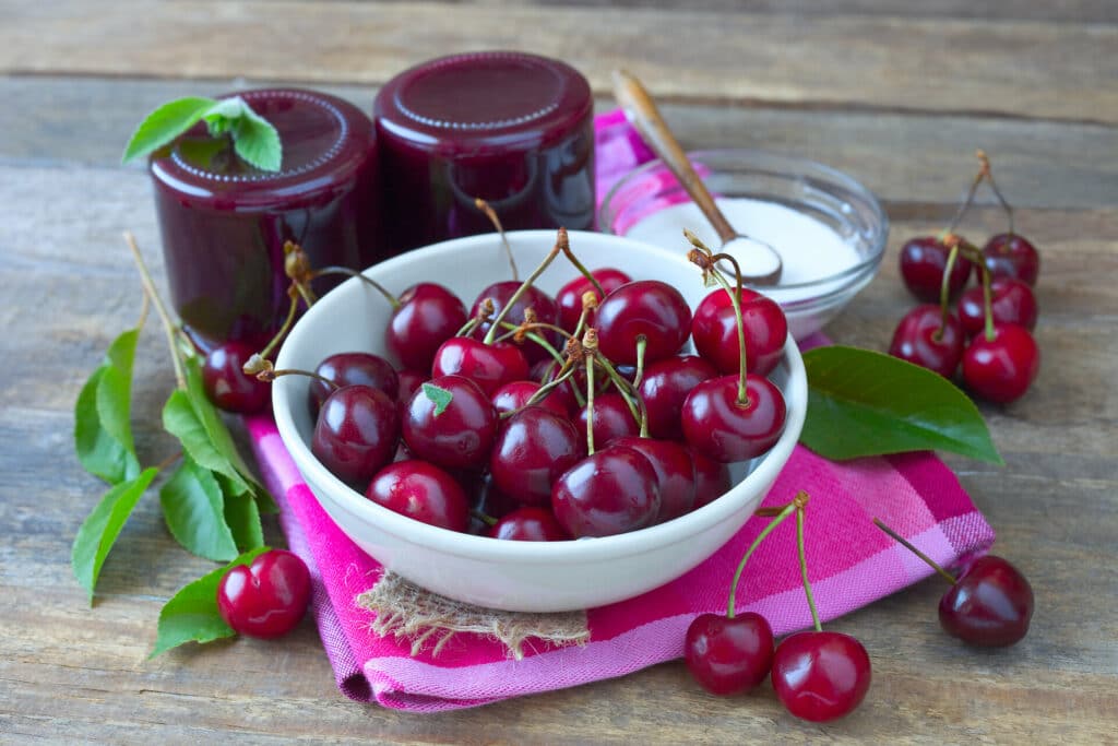 weißes Porzellanschälchen mit dunkelroten Kirschen auf pinkfarbenerm Küchentuch auf Holztisch daneben 2 auf den Kopf gestellte Marmeladengläser mit Kirchmarmelade und ein Schälchen mit Zucker auf Holztisch