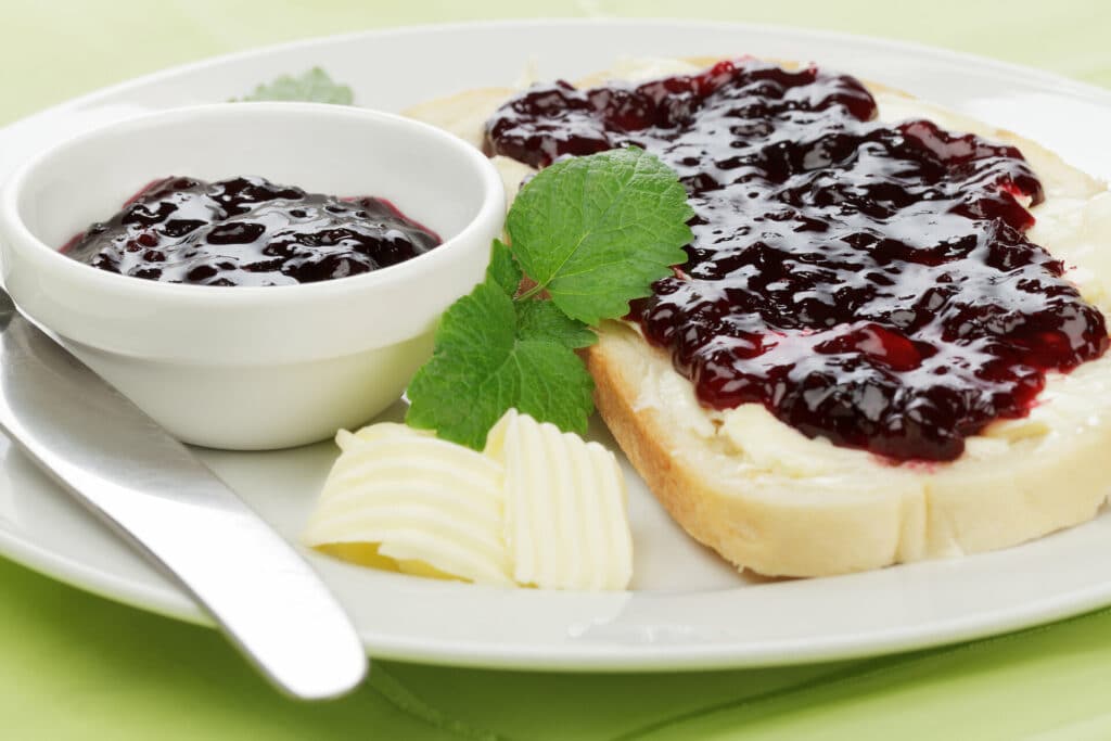 Weißbrot mit Butter und Kirschmarmelade auf weißem Teller mit weißem Marmeladenschälchen, Butter und einem Messer und Melissenblätter dekoriert