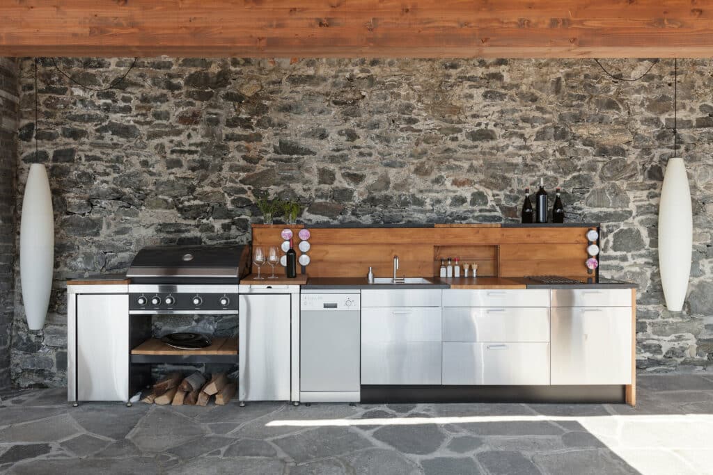 moderne Outdoorküche mit Grill und silberfarbenen Fronten und Holz vor Steinmauer