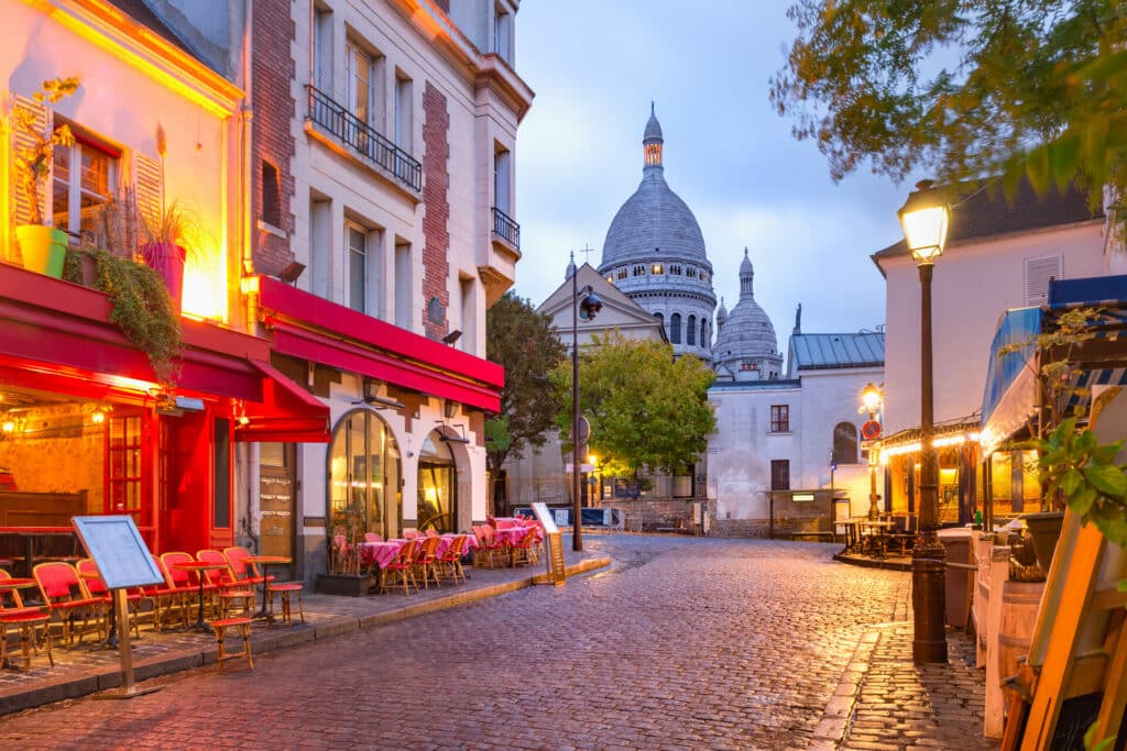 beleuchtete Cafes auf gepflasterter Straße in Montmatre Paris am Abend im Hintergrund Sacre Coeur.