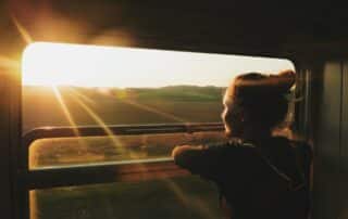 Junge Frau blickt aus dem Zugfenster in eine sonnige Landschaft