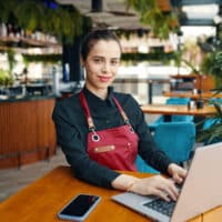 Restaurant Managerin mit roter Schürze und tippst auf Notebook, das auf einem Holztisch neben einem Mobiltelefon steht im einem Restaurant.