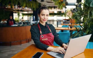 Restaurant Managerin mit roter Schürze und tippst auf Notebook, das auf einem Holztisch neben einem Mobiltelefon steht im einem Restaurant.