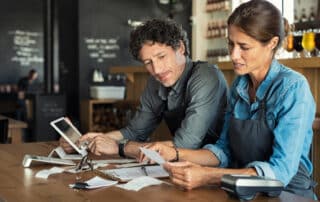 Mann und Frau sitzen an einem Restauranttisch mit Tablet und Rechenmaschine und vielen Belegen und machen die Buchhaltung