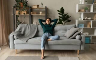 Junge dunkelhaarige Frau mit Jeans und dunkelgrünem Pulle sitzt auf einem grauen Sofa in ihrer Wohnung und genießt lächelnd Entspannung und Gemütlichkeit - Einrichtungstipps