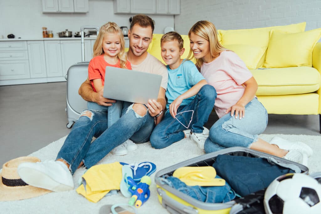 Familie mit einem Jungen und einem Mädchen sitzt zuhause auf dem Sofa mit Laptop und plant den Urlaub. Im Vordergrund ist ein geöffneter Koffer mit Kleidungsstücken, Flipflops, Wasserpistole und Fußball