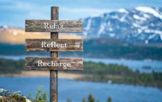 Holzschild vor Bergsee mit schneebedeckten Gipfeln mit der Aufschrift Reflect, Relax, Recharge - Wellnessurlaub